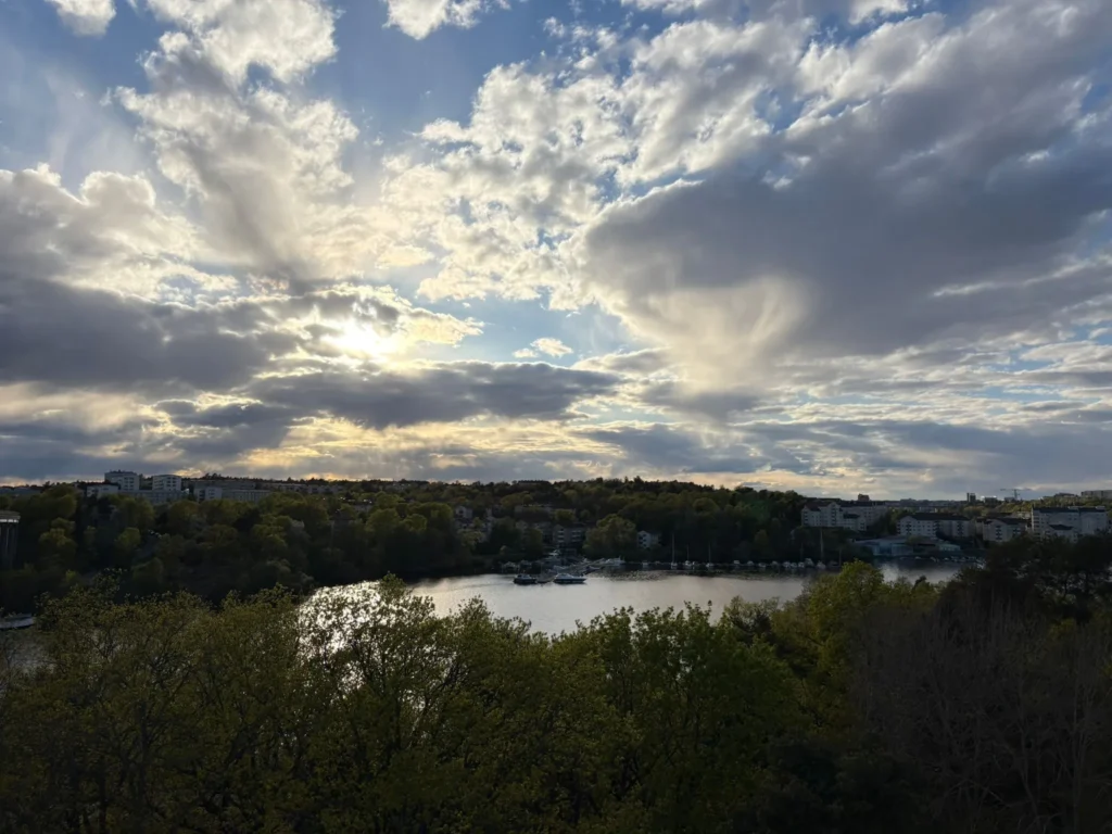 Ett vidvinkelfotografi taget från taket på Levantinsgatan 1, med utsikt över ett vattenområde mot ett bostadsområde i Alvik som ligger inbäddat i en lummig, grön skog. Scenen domineras av en dramatisk, vidsträckt himmel fylld av stora, texturerade vita och grå moln. Solen är delvis skymd av molnen, vilket skapar en "solstråle-effekt" där ljuskäglor strålar utåt och reflekteras med glans i vattenytan nedanför. I mellanplan syns en lugn vik eller ett vattendrag, med flera små båtar förtöjda längs den bortre strandlinjen. Den motsatta sidan består av ett tätt krön av gröna träd, där olika flerbostadshus och lägenhetskvarter tittar fram genom grönskan. Förgrunden är fylld av mörka silhuetter från trädtoppar, vilket ramar in utsikten och förstärker känslan av höjd.