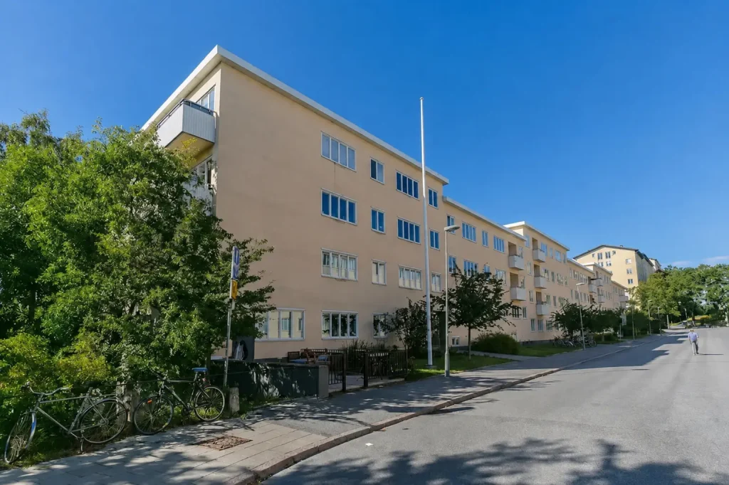 Ett beige bostadshus i funkisstil längs en asfalterad gata under en klarblå himmel. Framför huset syns en tom flaggstång, gröna träd och parkerade cyklar till vänster.